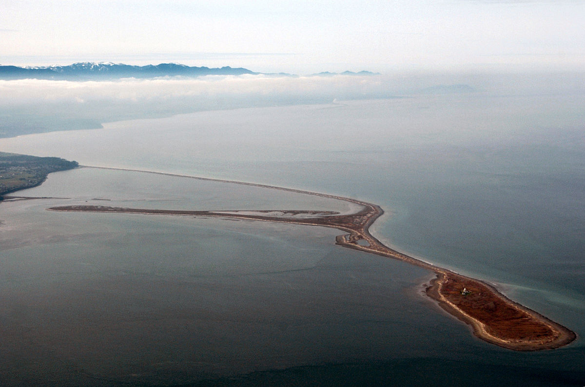 Aerial view of a body of water with a long, narrow island or peninsula extending into it, with mountains in the background and clouds or fog rolling in.